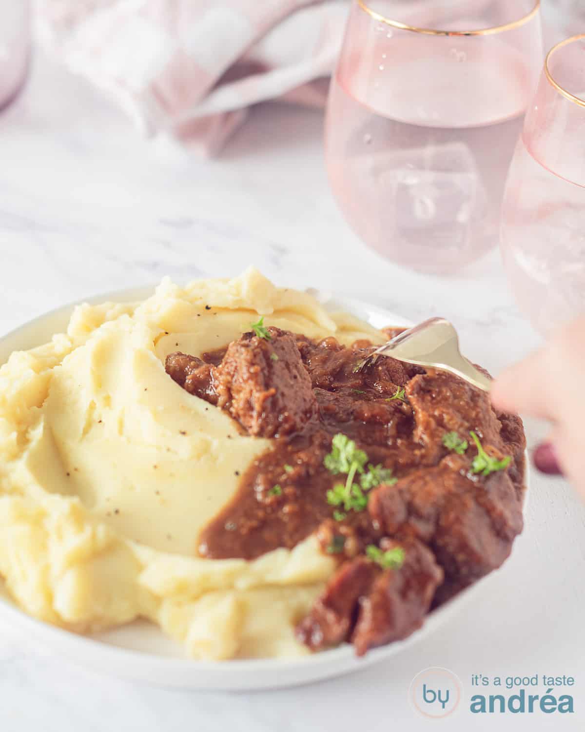 Een wit bord met een stoofschotel met speculaas en appelstroop en daarnaast aardappelpuree. Een vork neemt een hap. Een wit gemarmerde ondergrond. In de achtergrond twee roze glazen en een roze witte theedoek.