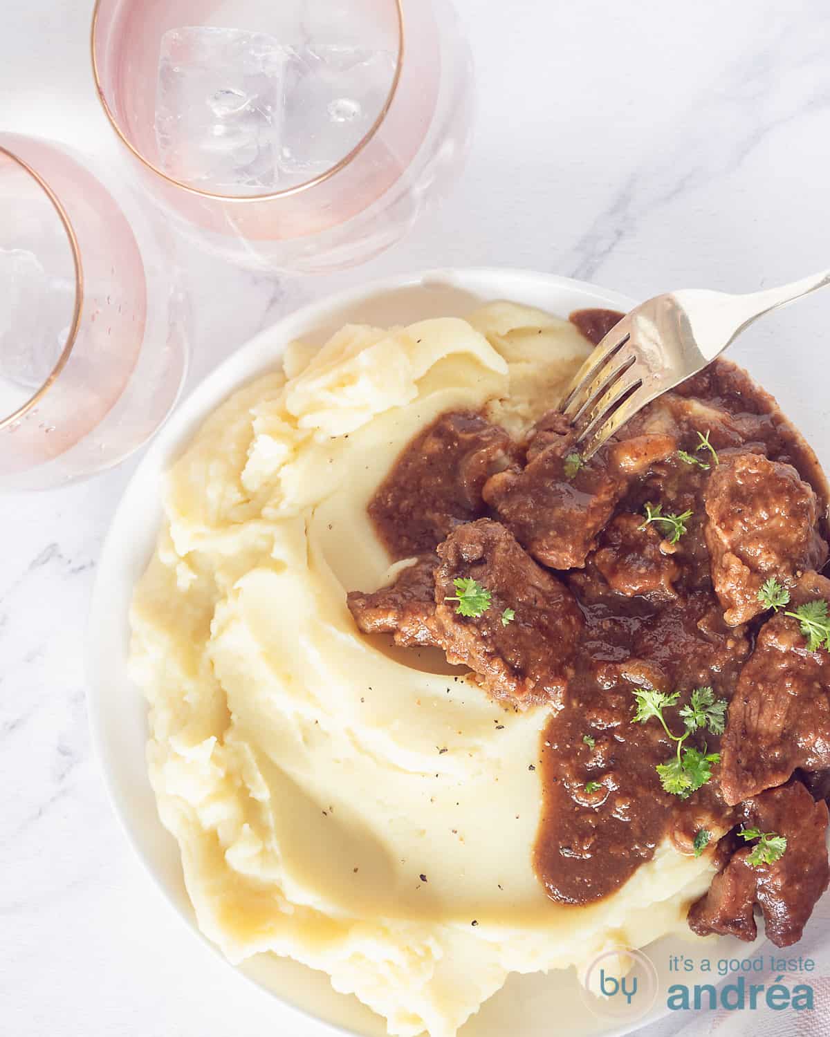 Een foto van bovenaf van een wit bord met aardappelpureee en een stoofpotje met speculaas. Op een wit gemarmerde ondergrond. Linksbovenin twee roze glazen.