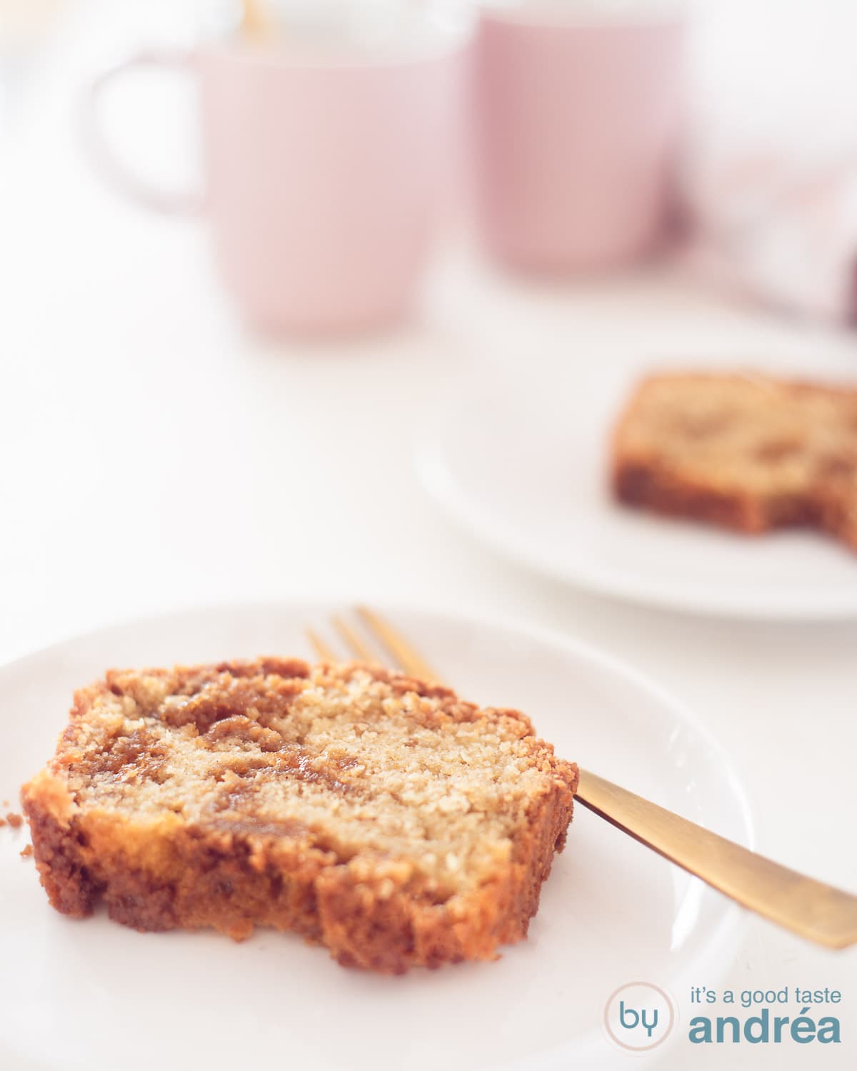 Twee plakjes cake op een wit bordje achter elkaar op een witte ondergrond. In de achtergrond twee theemokken.