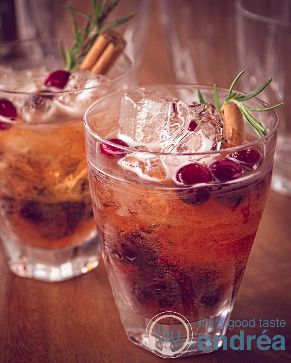 Thanksgiving cranberry cocktail twee stuks schuin achter elkaar op een houten tafel. Met een stokje kaneel, cranberries en een takje rozemarijn.