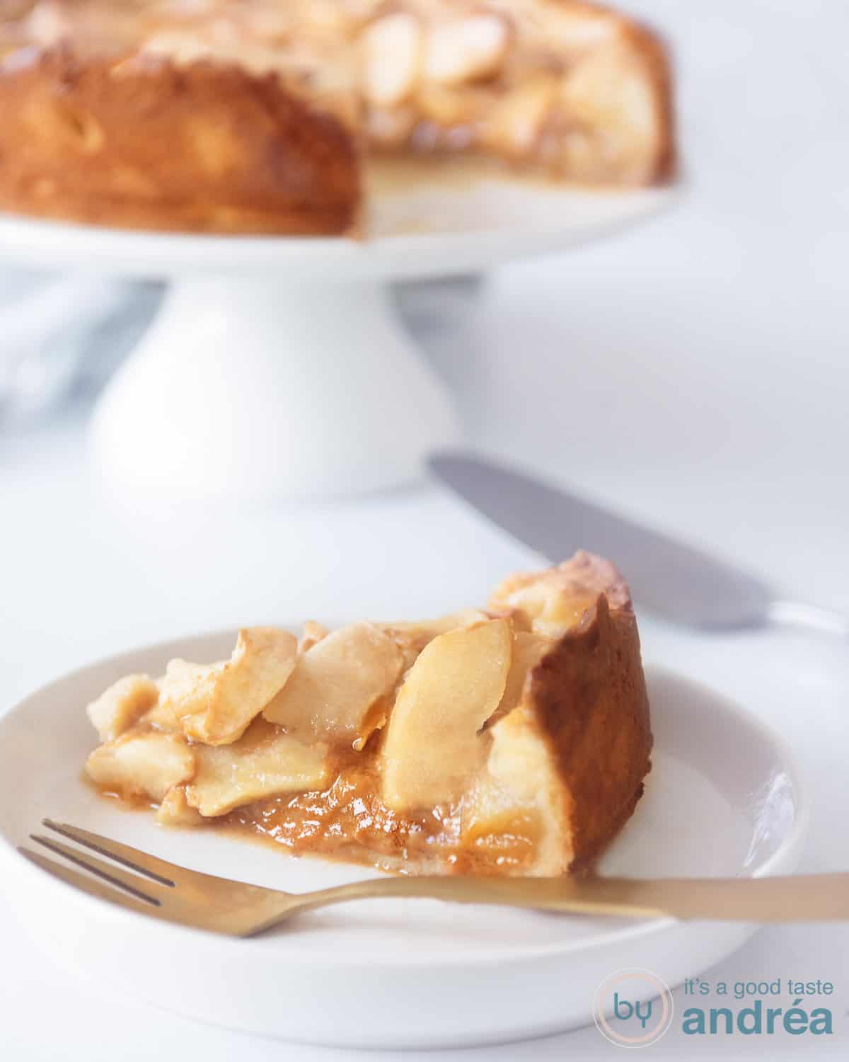 Een stukje appeltaart op een wit bord gevuld met karamel. In de achtergrond een taartstandaard met de rest van de taart