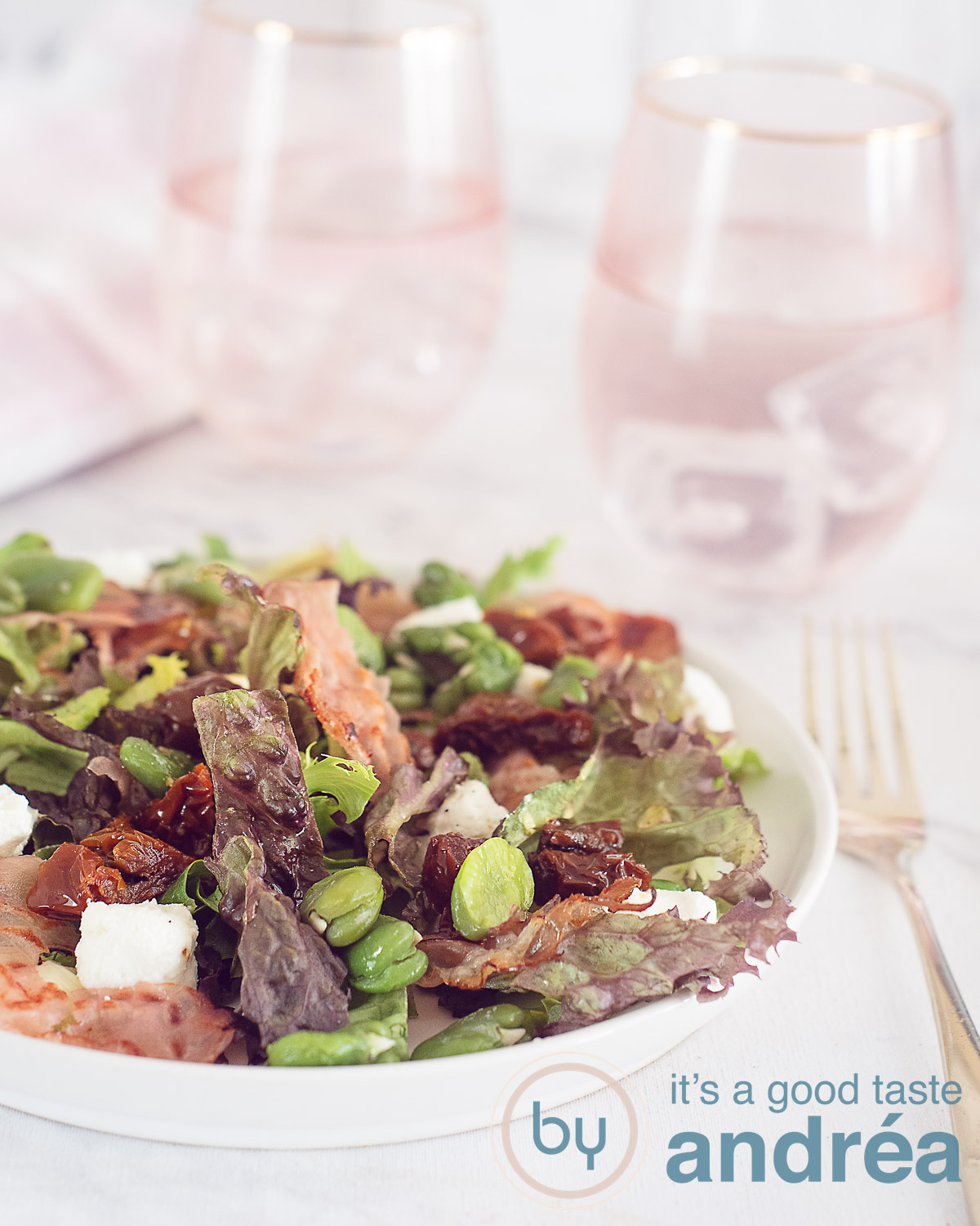 Een hoogtefoto met een deel van een wit bord gevuld met een tuinbonensalade met geitenkaas en bacon. Een witte ondergrond. In de achtergrond twee roze glazen gevuld met water.