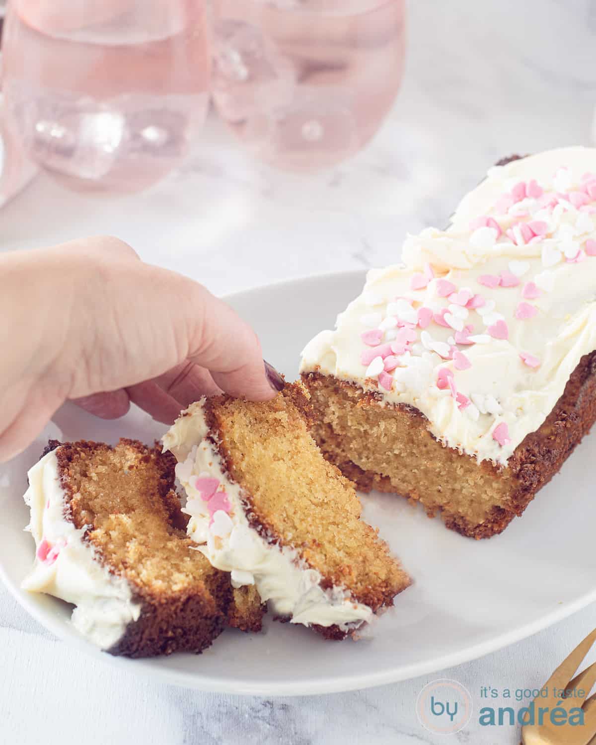 witte chocolade cake met een topping van witte chocolade pasta en sprinkles op een wit bord. Een hand pakt een plakje