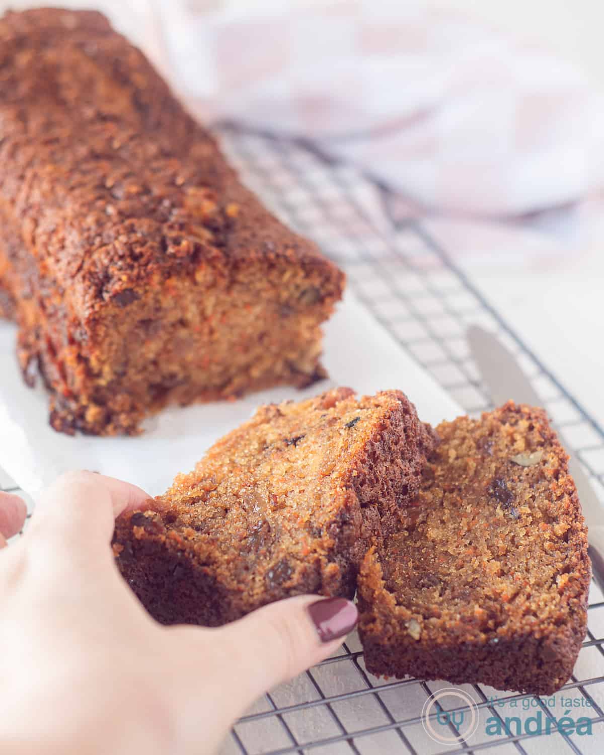 Wortelcake met rozijnen en noten op een rek, waar twee plakjes vanaf zijn gesneden. Een hand pakt een plakje.