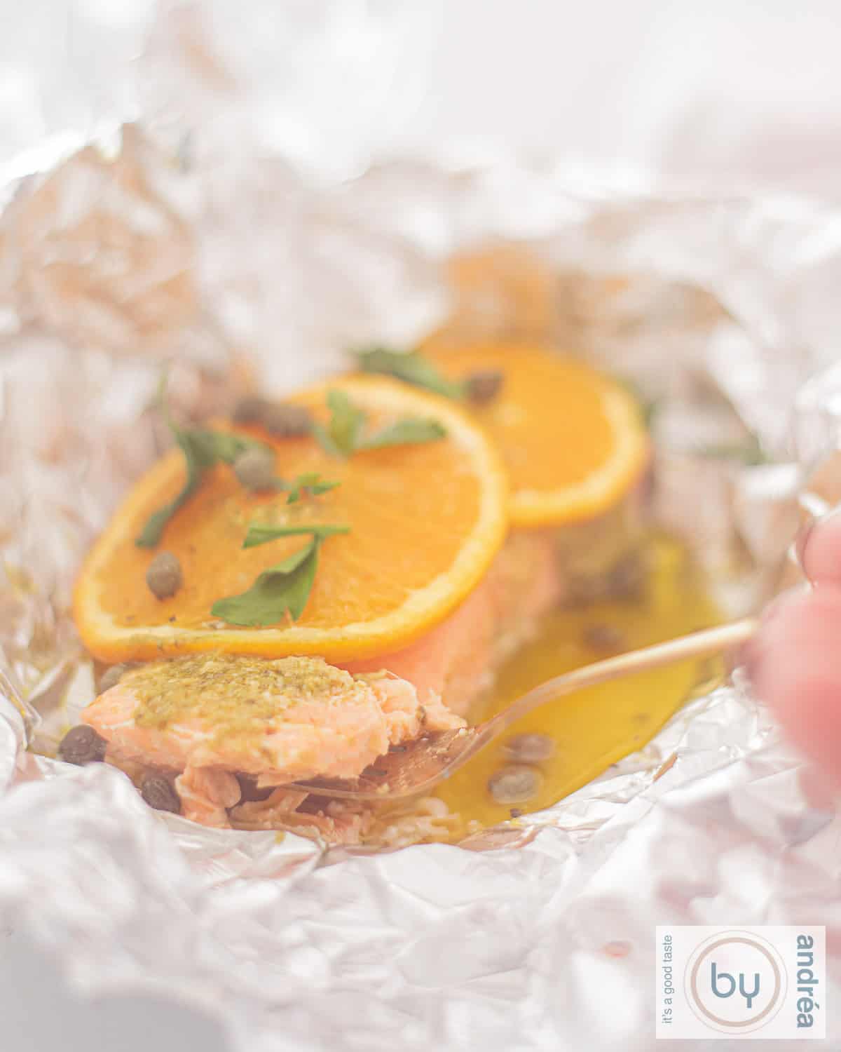 aluminiumfolie daarop zalm met schijfjes sinaasappel, gesmolten boter, pesto en kappertjes. Een vork pakt een hap