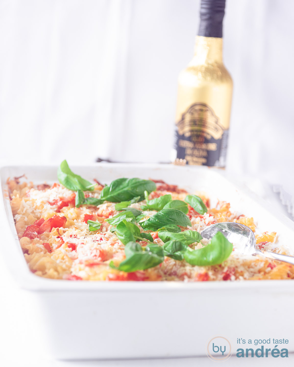 pasta caprese ovenschotel in een witte ovenschotel op een witte ondergrond. Een gouden fles olijfolie in de achtergrond.