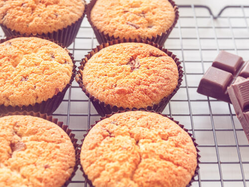 Een vierkante foto met 6 cupcakes met chocoladestukjes op een rek. Chocola ligt er rechts naast.