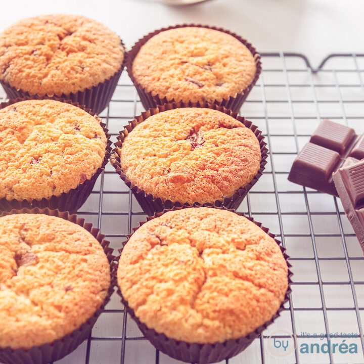 Een vierkante foto met 6 cupcakes met chocoladestukjes op een rek. Chocola ligt er rechts naast.