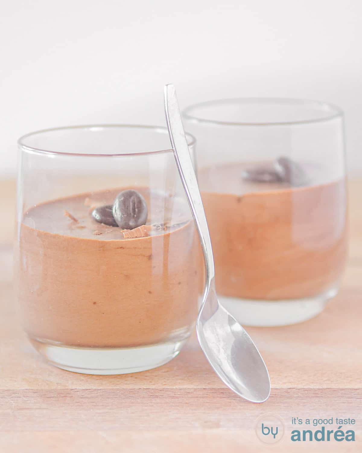 2 glaasjes met Eenvoudige chocolade mousse op een houten plank. Een lepel staat tegen de glaasjes aan.