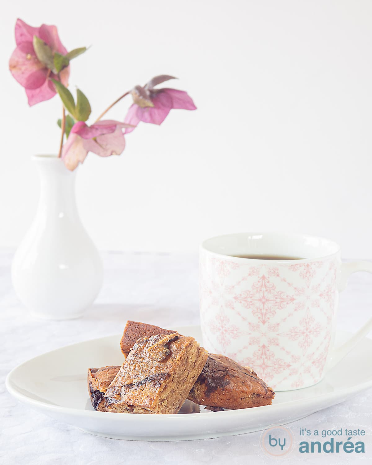 Een wit bord met honing blondies met een roze witte mok gevuld met koffie. Op een witte ondergrond. Links achter een vaas met bloemen.