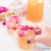 Een vierkante foto met twee glazen ijsthee schuin achterelkaar. Decoratie van roos en framboos en limoen. Een hand pakt het voorste glas.