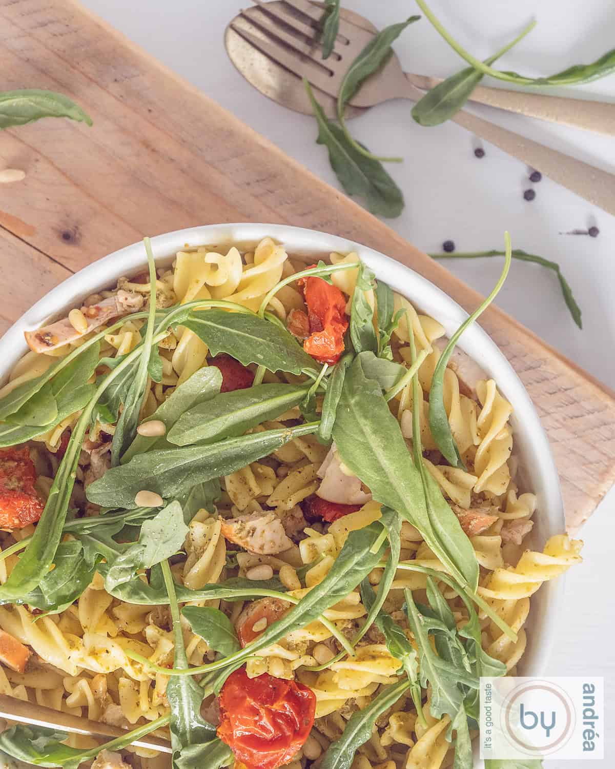 Een foto van bovenaf van pastasalade met gerookte kip, rucola en pesto op een houten plank.
