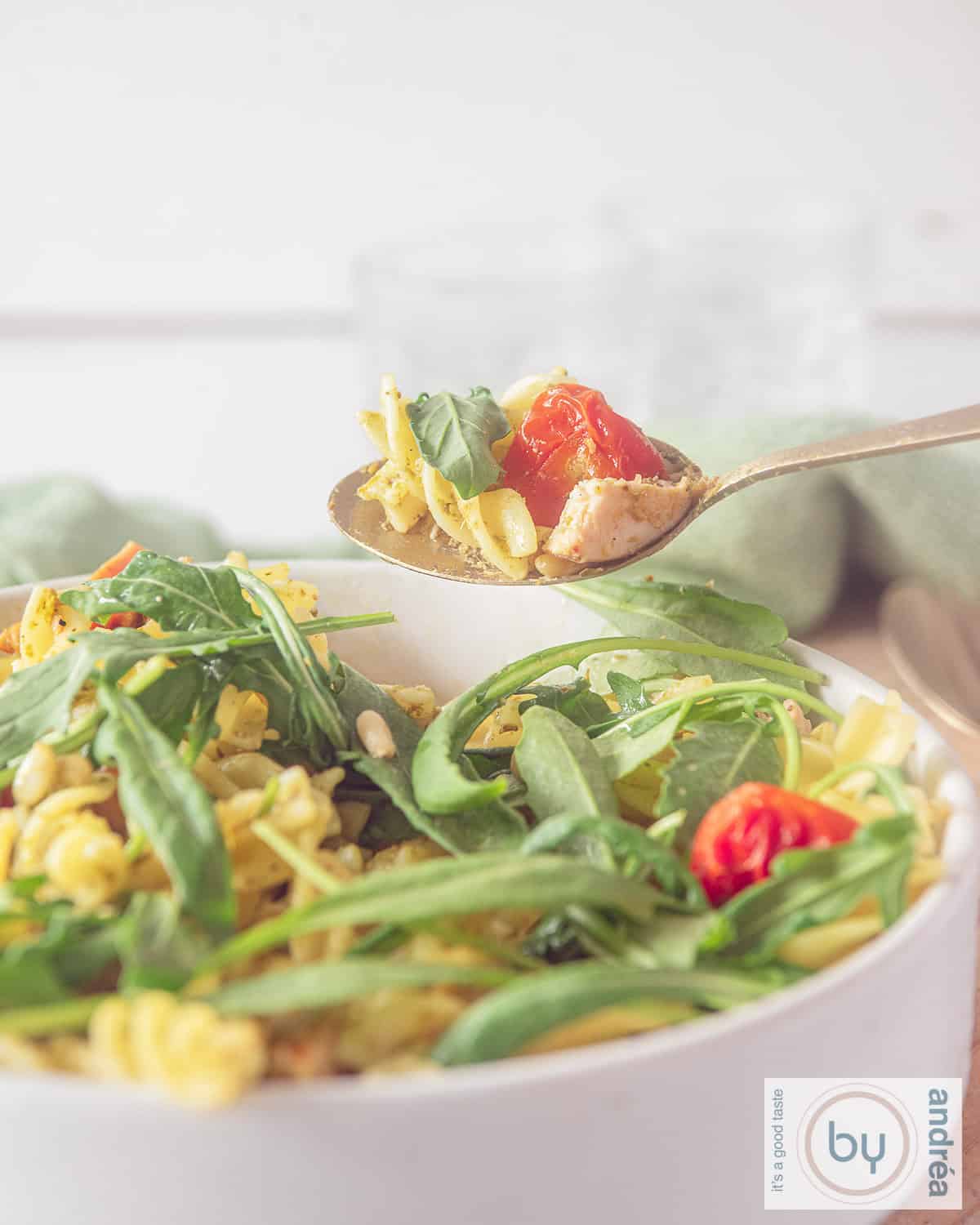 Een witte schaal met pastasalade met gerookte kip, rucola en pesto. Een lepel neemt een hap uit de schaal.