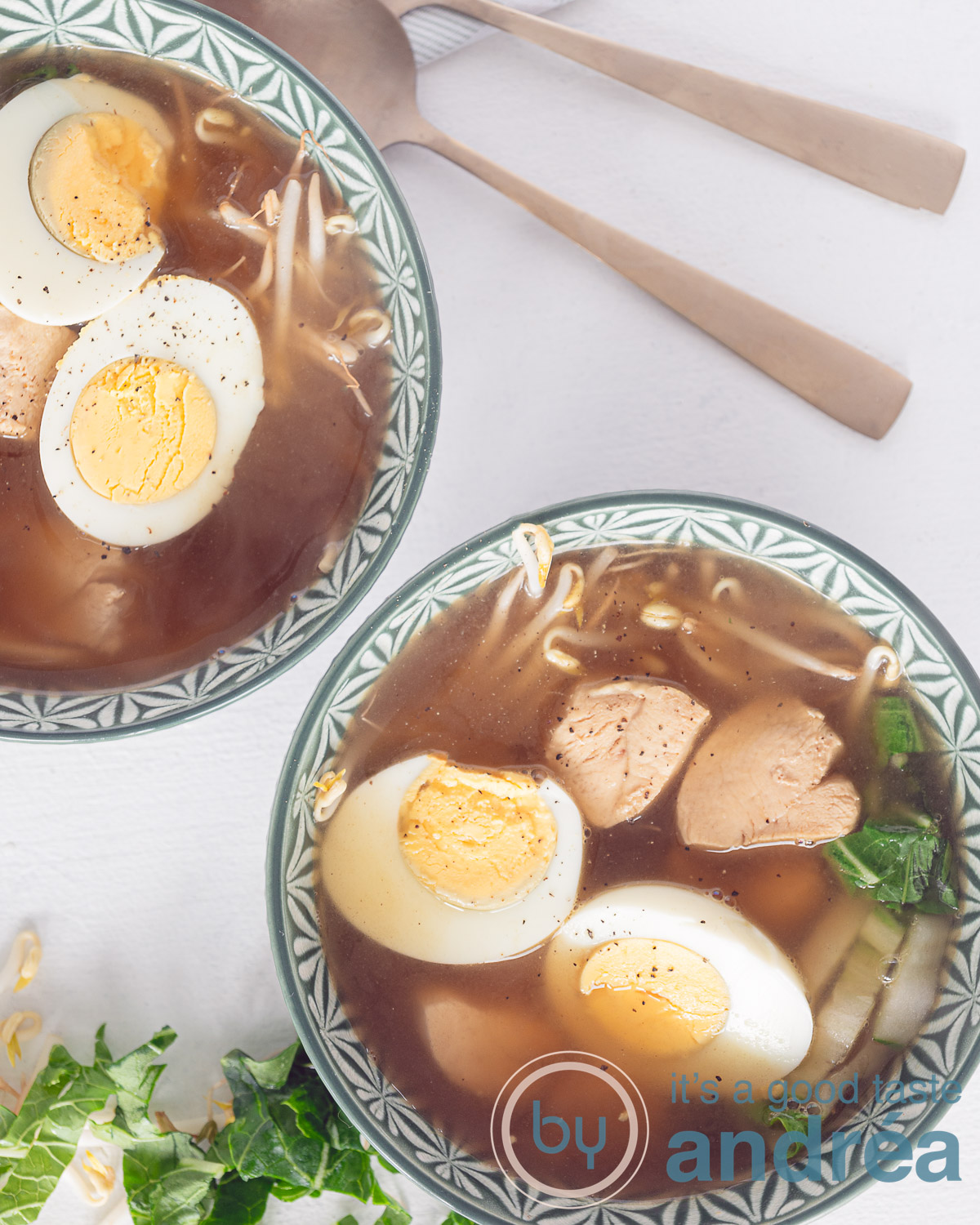 Een foto van bovenaf van twee groene kommen gevuld met Ramen noedelsoep met kip, eierenk tauge en paksoi. twee lepels liggen erbovenin. Een witte ondergrond.