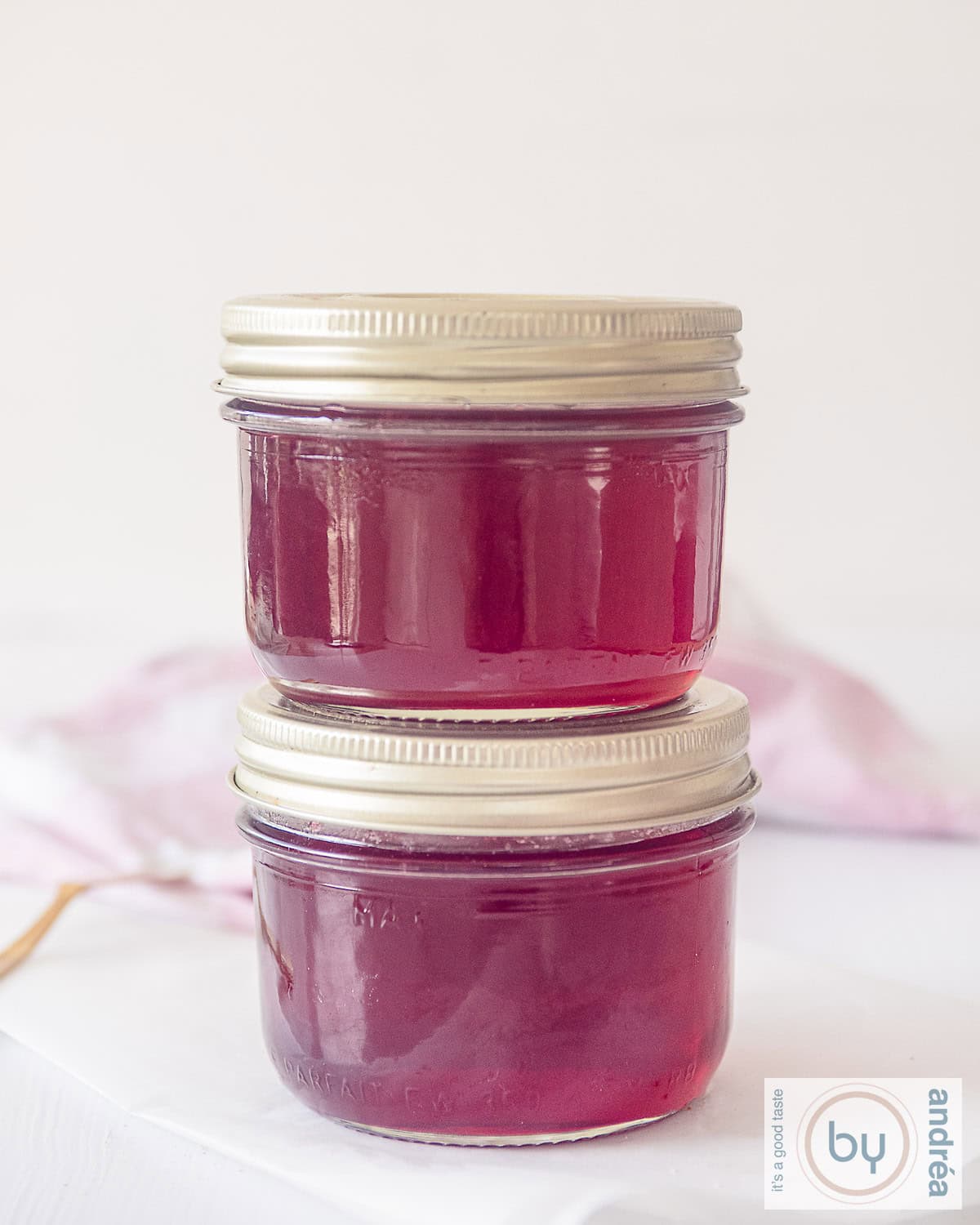 Twee potten rode bessenjam in een glazen pot met gouden deksel op elkaar. Een witte ondergrond met in de achtergrond een roze witte theedoek.