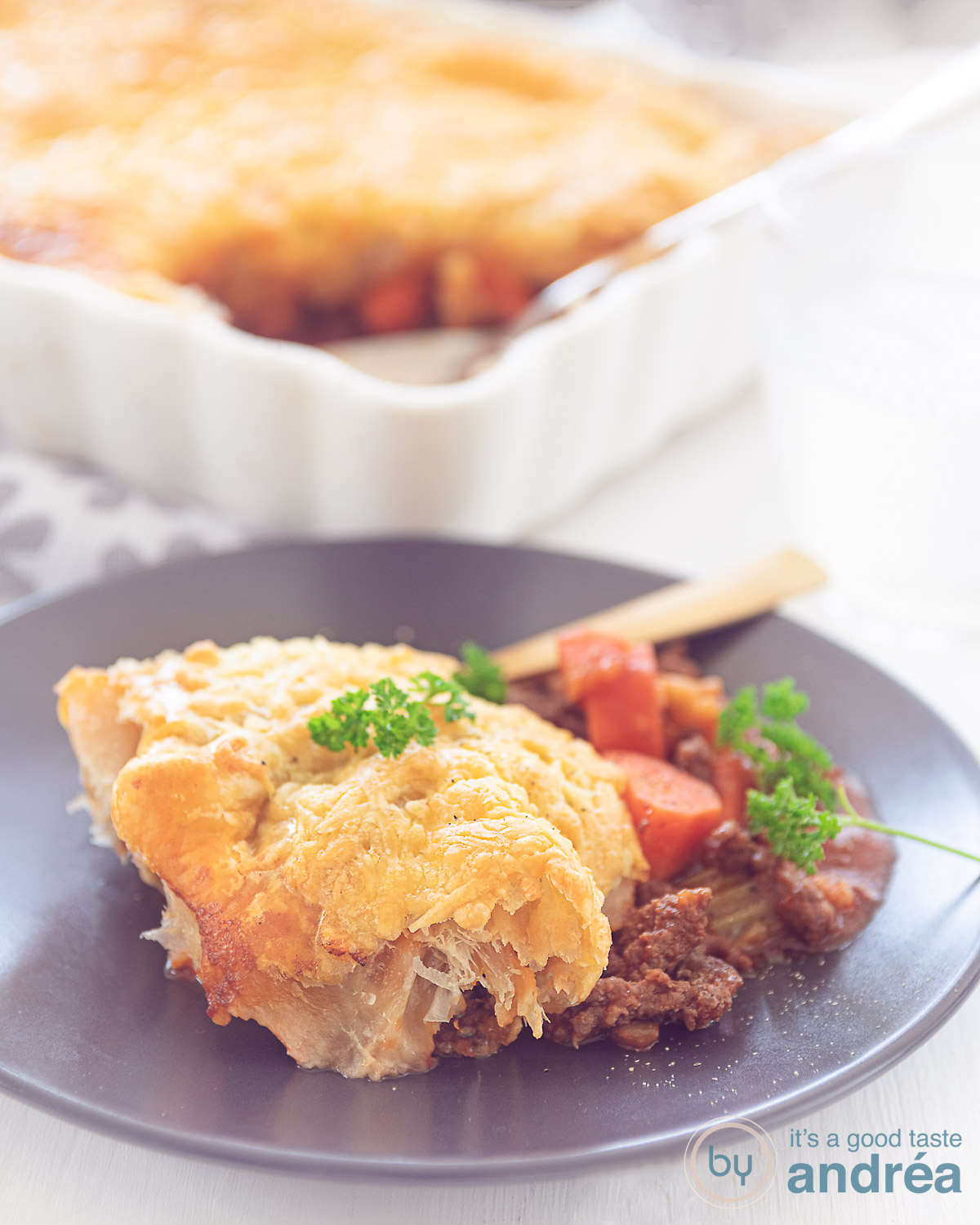 Een stuk shepherd's pie op een grijs bord. In de achtergrond de rest van de ovenschaal. Een witte ondergrond.