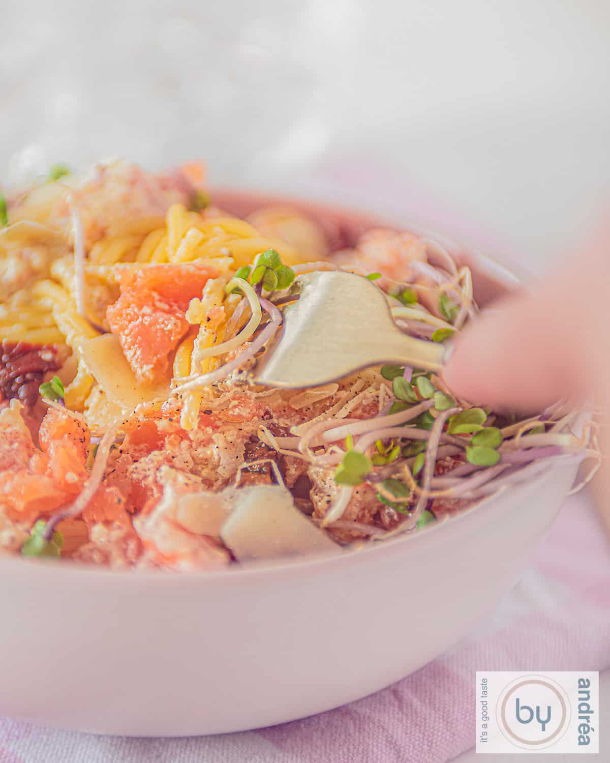 Een deel van een roze kom gevuld met spaghetti, gerookte zalm, zongedroogde tomaatje en creme fraiche. Een vork prikt in de spaghetti