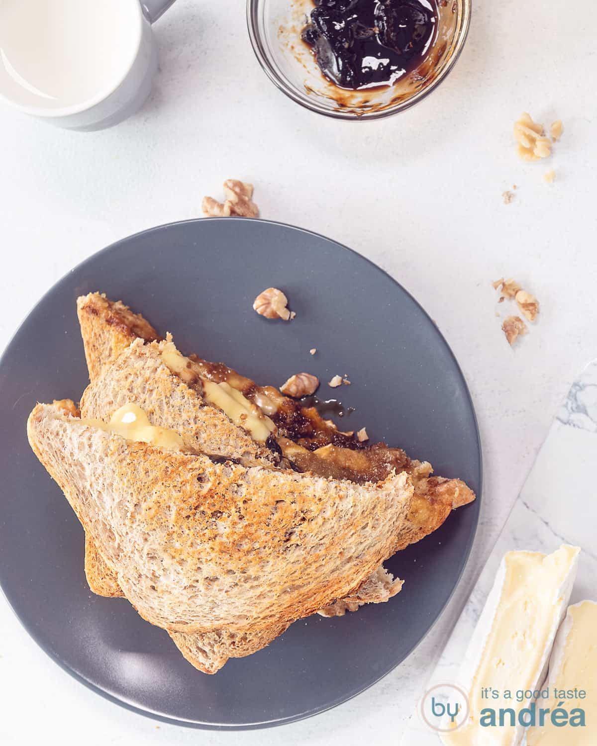 Een foto van bovenaf van een tosti met camembert, appelstroop en noten op een witte ondergrond. Rechts wat camembert, bovenin appelstroop en een beker melk.