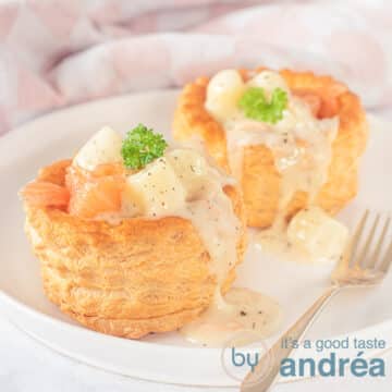 Een vierkante foto met een wit bord met twee bakjes gevuld met gerookte zalm en asperge ragout wat over het bakje heen druipt. Een gouden vork ligt erbij.