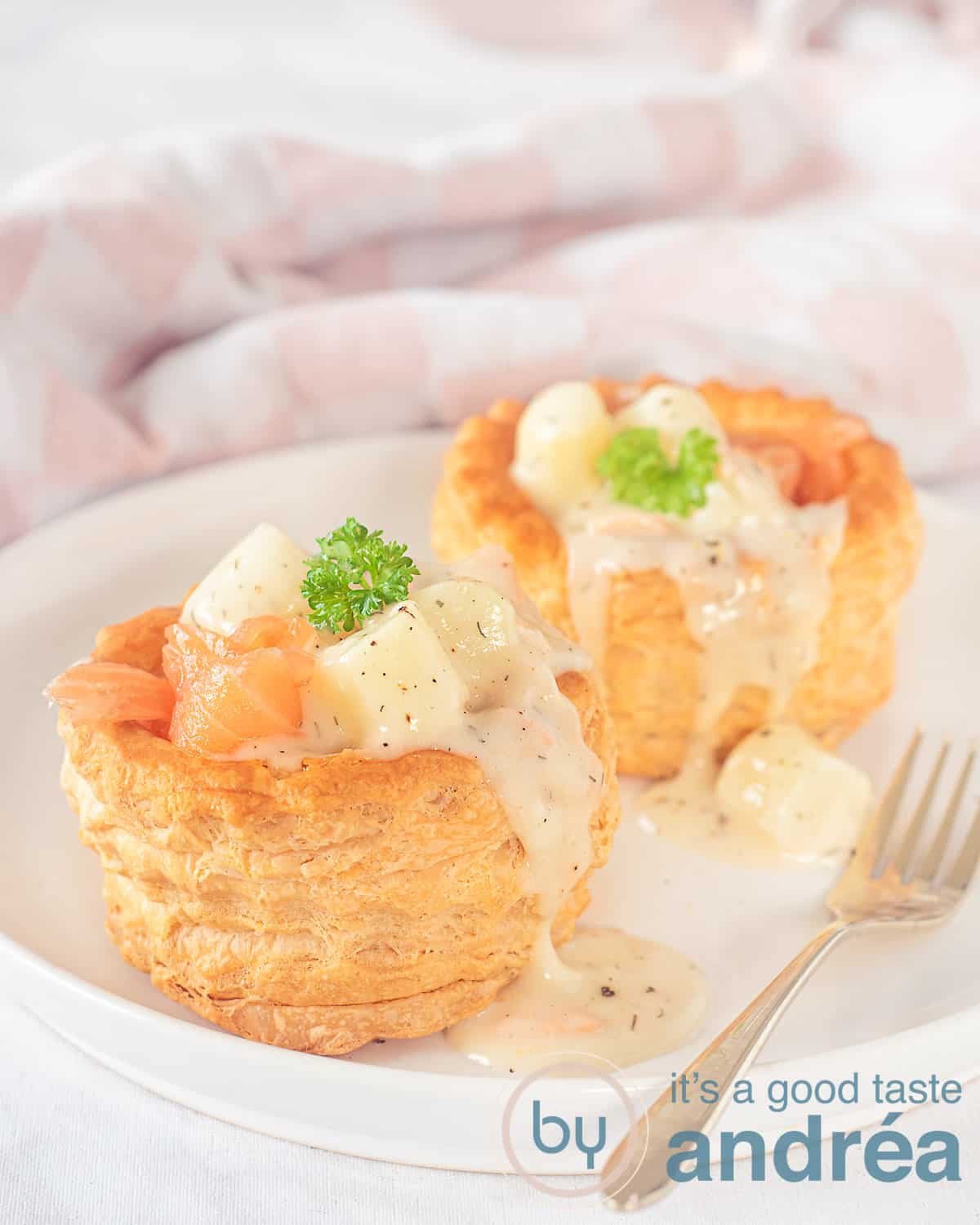 Een hoogtefoto van twee bladerdeegbakjes gevuld met asperge met gerookte zalm ragout op een wit bord. Een gouden vork ligt erlangs. In de achtergrond een wit roze geblokte theedoek