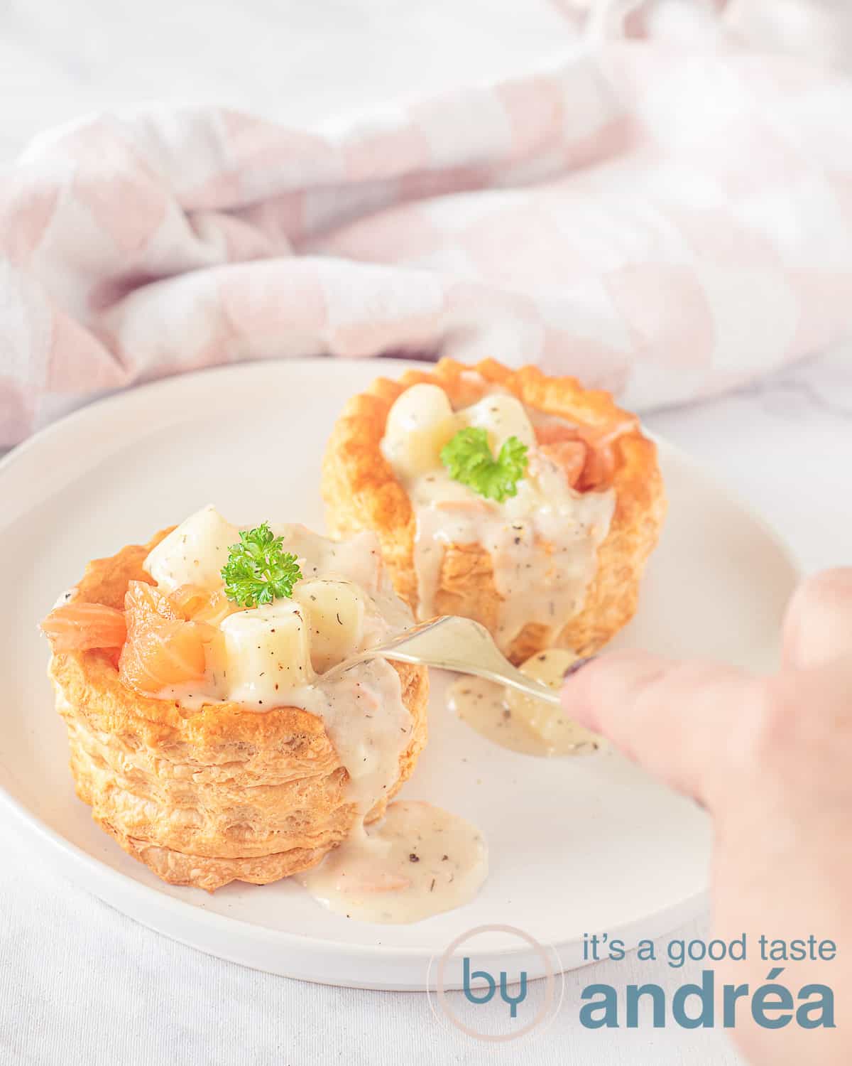 Een hoogtefoto met een wit bord met twee ragoutbakjes met aspergeragout en zalm. Een vork prikt in de ragout. In de achtergrond een roze witte geblokte theedoek.