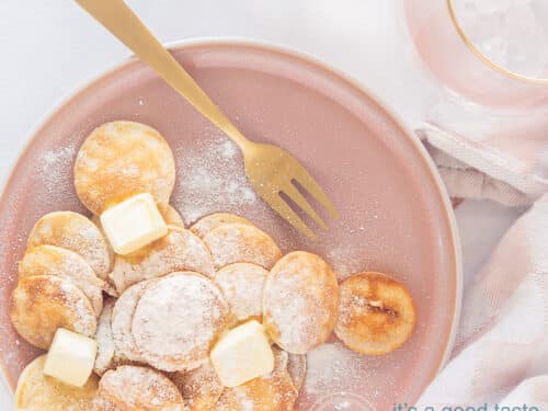Een vierkante foto met een roze bord met een stapel mini pannenkoekjes, boter en poedersuiker een gouden vork en een witte ondergrond