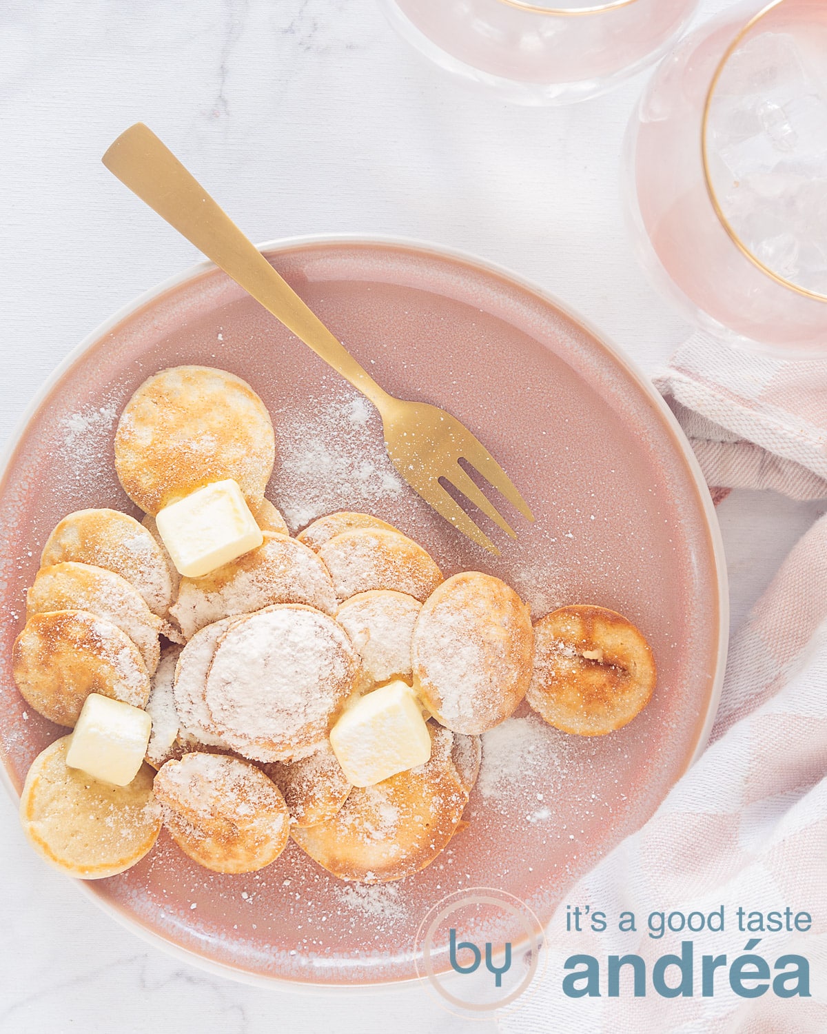 Een foto van bovenaf, in de hoogte, van een roze bord met poffertjes, poedersuiker en boter. Een gouden vork ligt erboven. Een witte ondergrond. Rechts bovenin een deel van twee roze glazen.