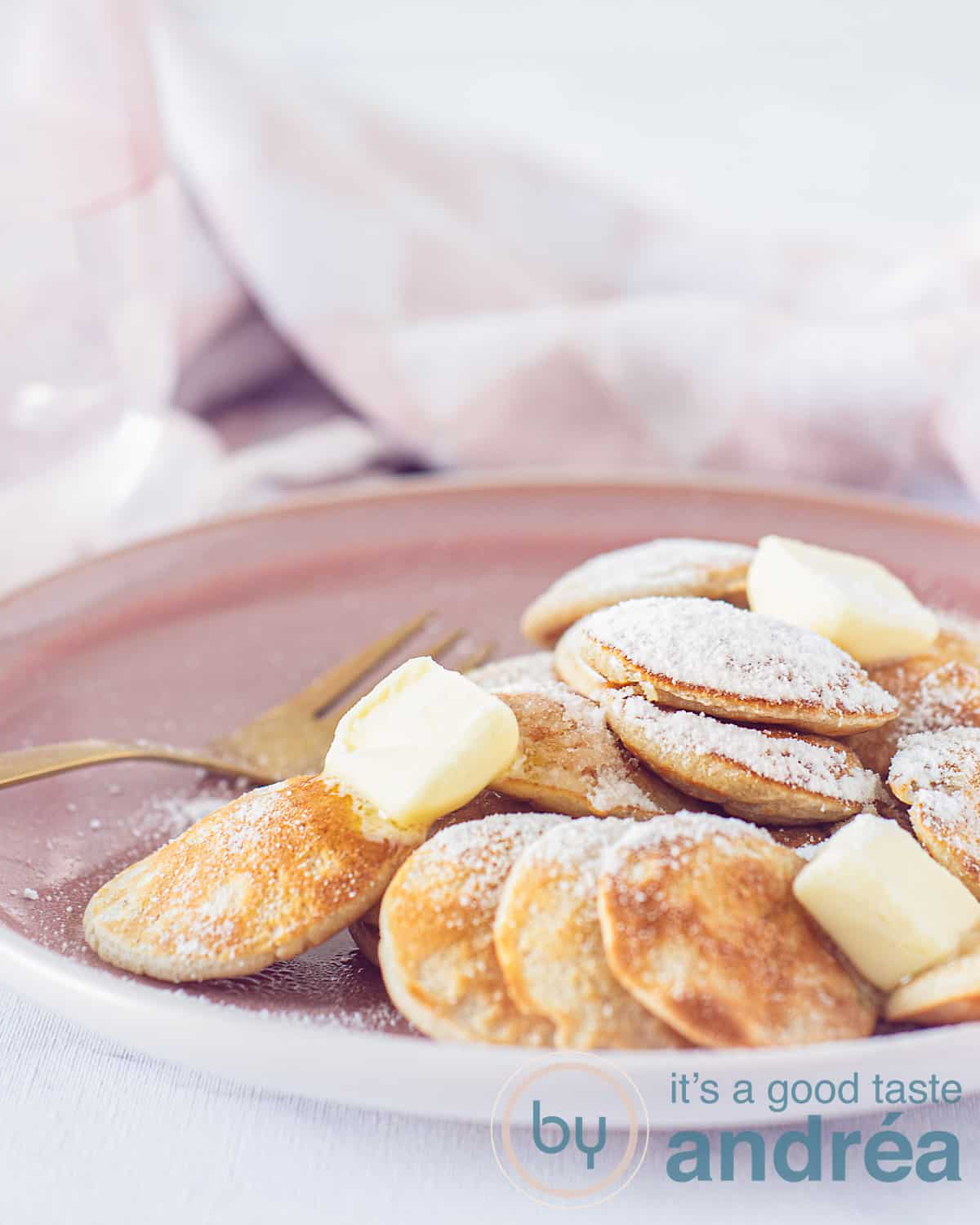 Een hoogtefoto met een deel van een roze bord met een stapel poffertjes, boter en poedersuiker. In de achtergrond een roze witte theedoek.