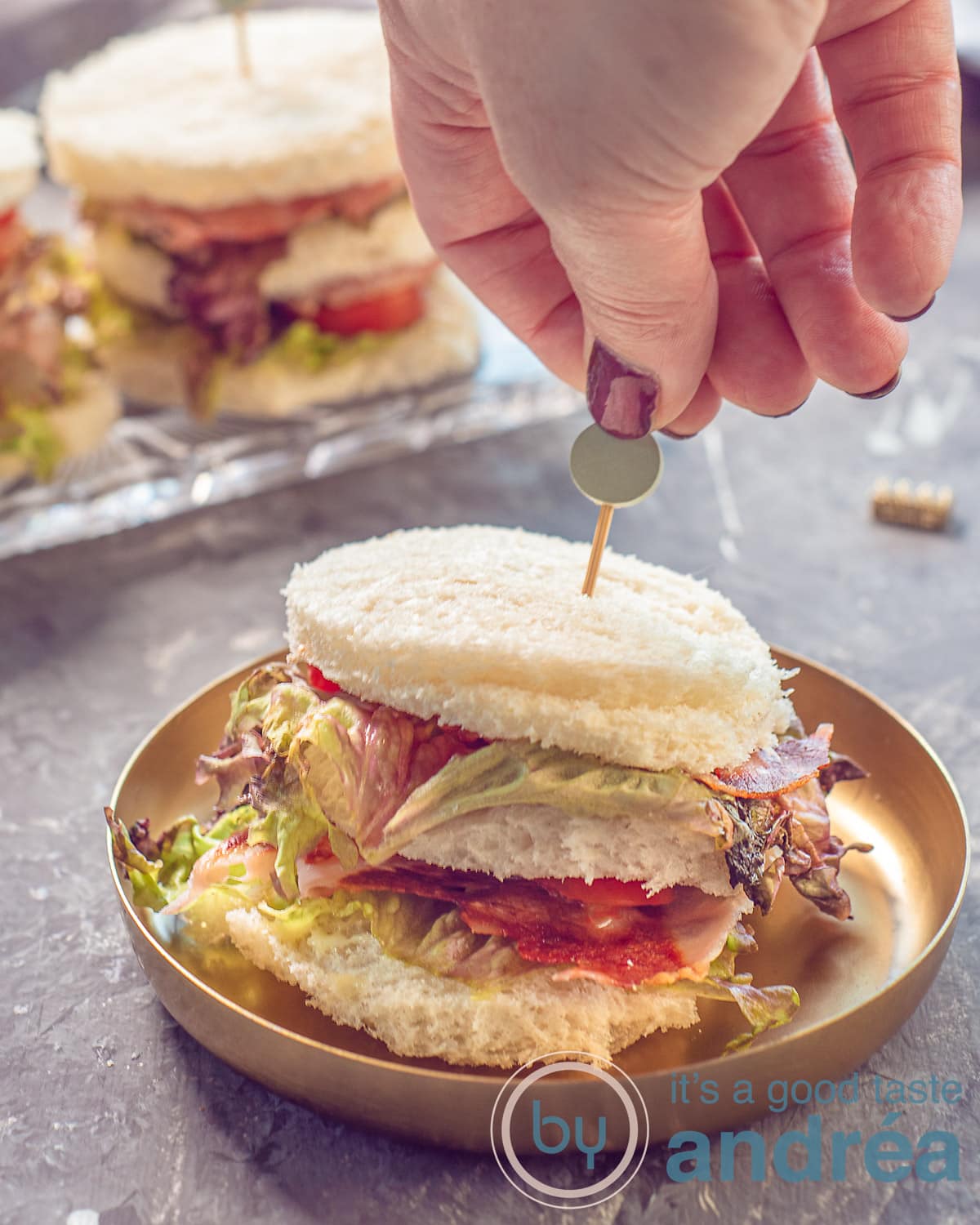 Een foto in de hoogte met een gouden bord met daarop een BLT mini Sandwich. Een hand prikt er een tapas prikker in. In de achtergrond meer sandwiches