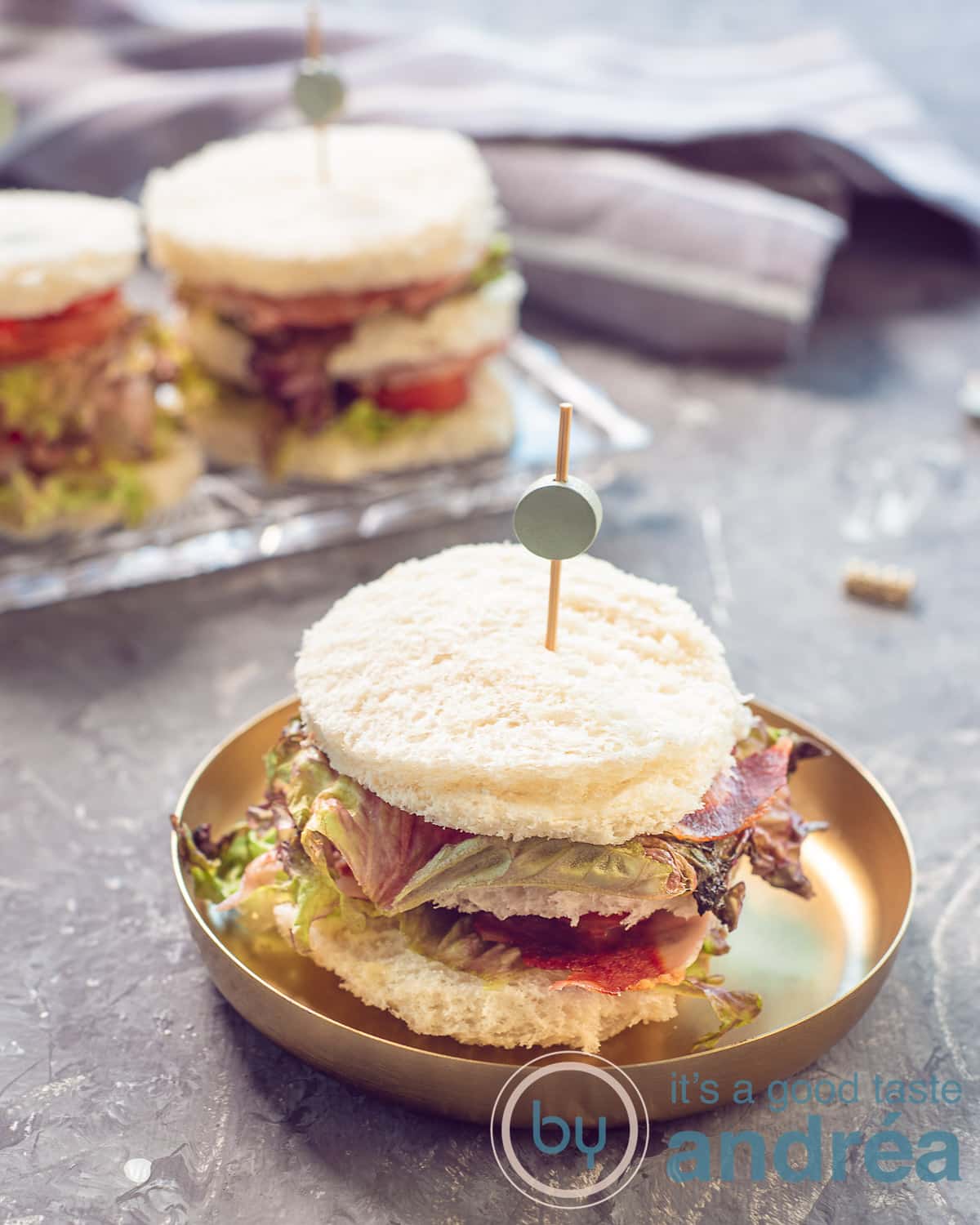 Een foto in de hoogte van een schaaltje van goud met een bacon sla en tomaat sandwich met een prikker erin. Een grijze ondergrond. In de achtergrond nog meer BLT sandwiches.