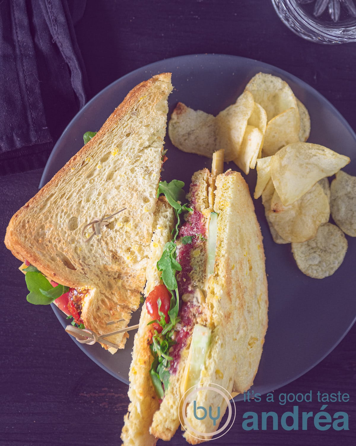 Een foto van bovenaf van twee sandwiches gevuld met rundercarpaccio, oude kaas en groenten met een heerlijke pestomayonaise. Wat chips erlangs. Een donkere ondergrond.