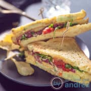 Een vierkante foto met twee driehoekjes van een clubsandwich met carpaccio, kaas en groenten op een grijze ondergrond.