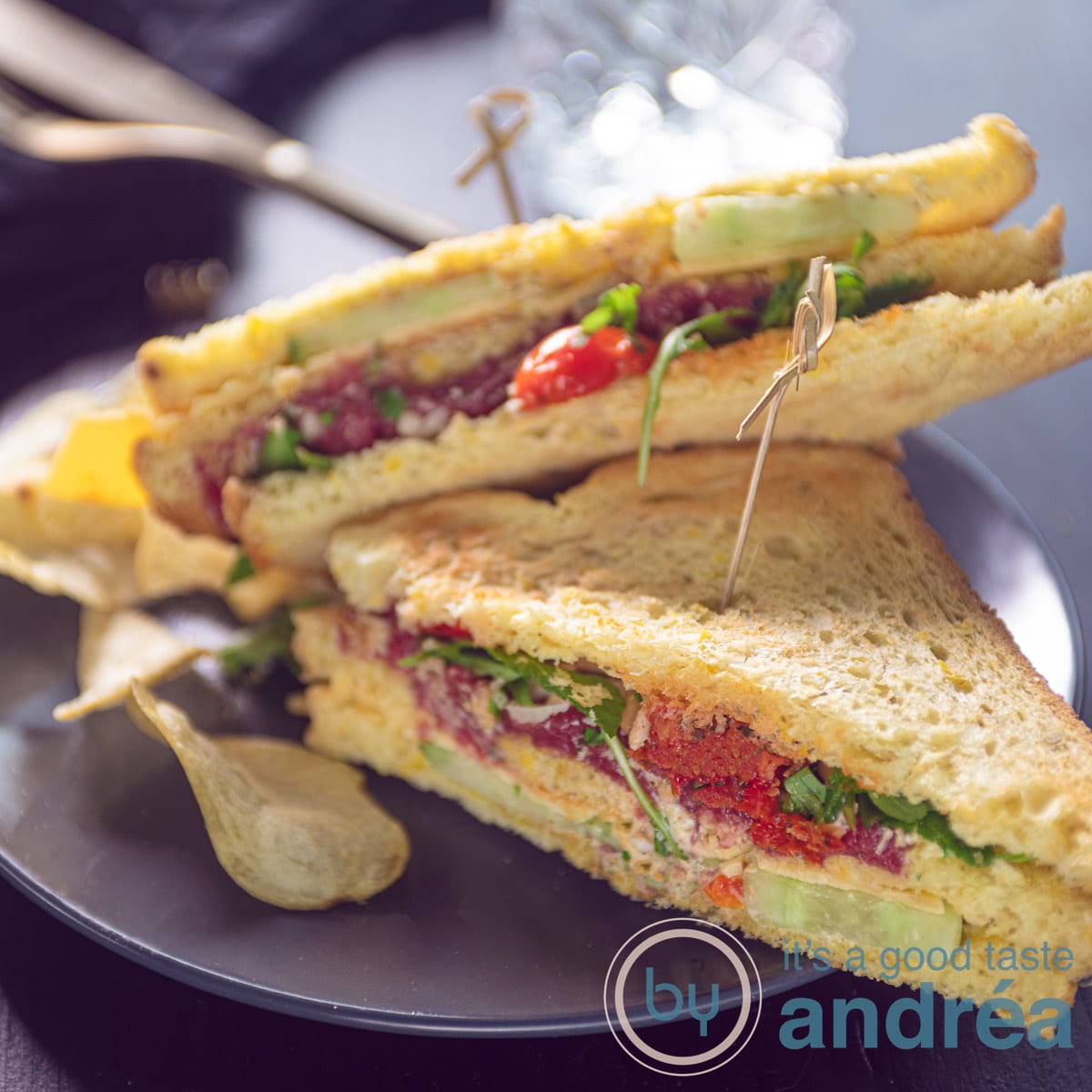 Een vierkante foto met twee driehoekjes van een clubsandwich met carpaccio, kaas en groenten op een grijze ondergrond.