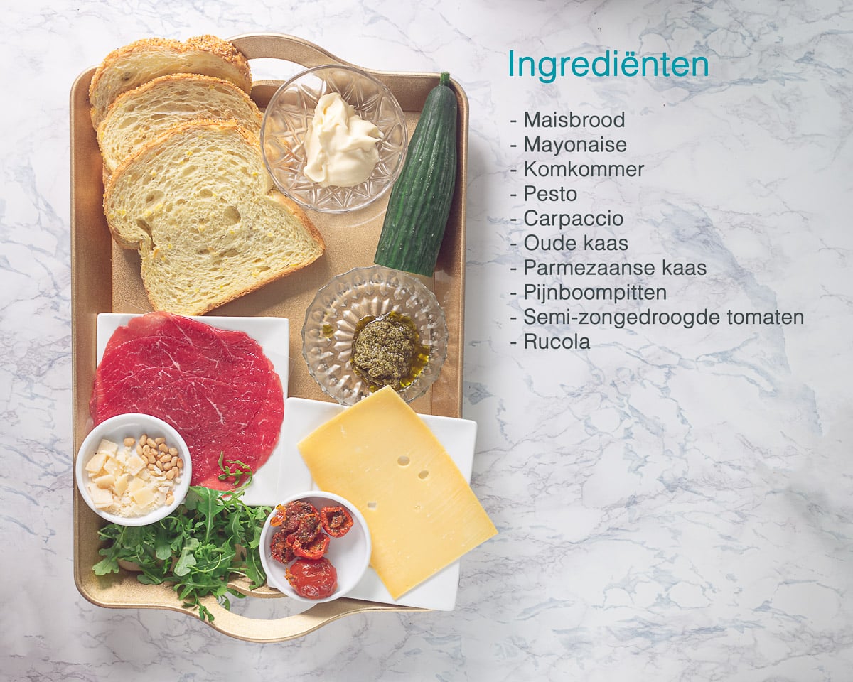 Ingrediënten Carpaccio Clubsandwich op een gouden dienblad. Er naast op een wit marmeren ondergrond een opsomming van de ingrediënten die nodig zijn.