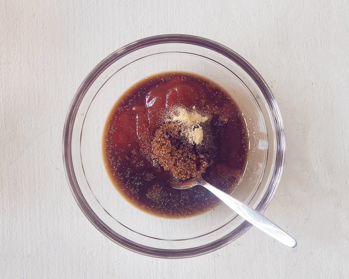 Alle ingrediënten voor de tonkatsu saus in een glazen kommetje met een lepel. Een witte ondergrond.