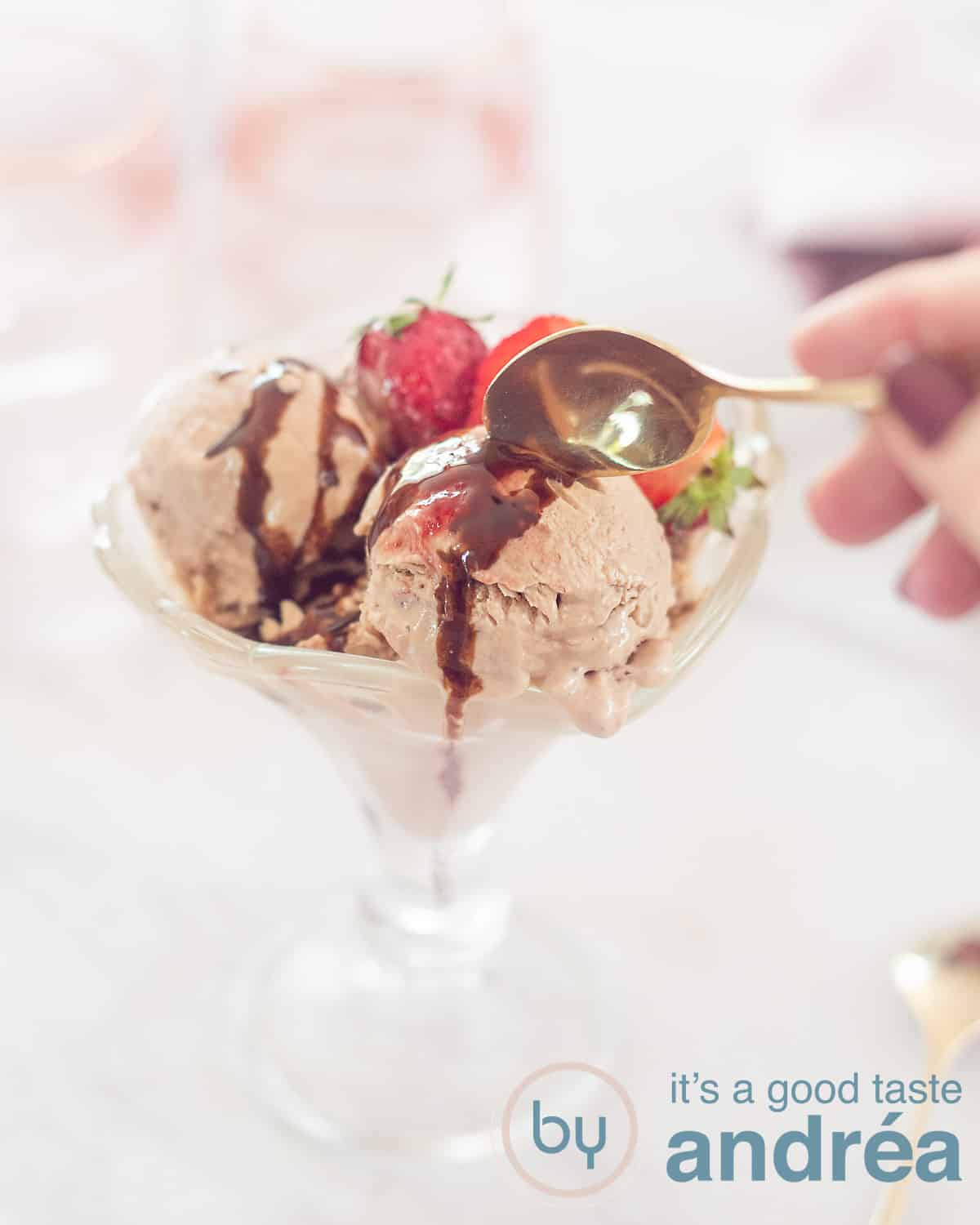 Een hoogtefoto met een glazen coupe gevuld met drie bolletjes yoghurtijs met chocola en aardbeien. Een gouden lepel pakt een hap. Een witte ondergrond.