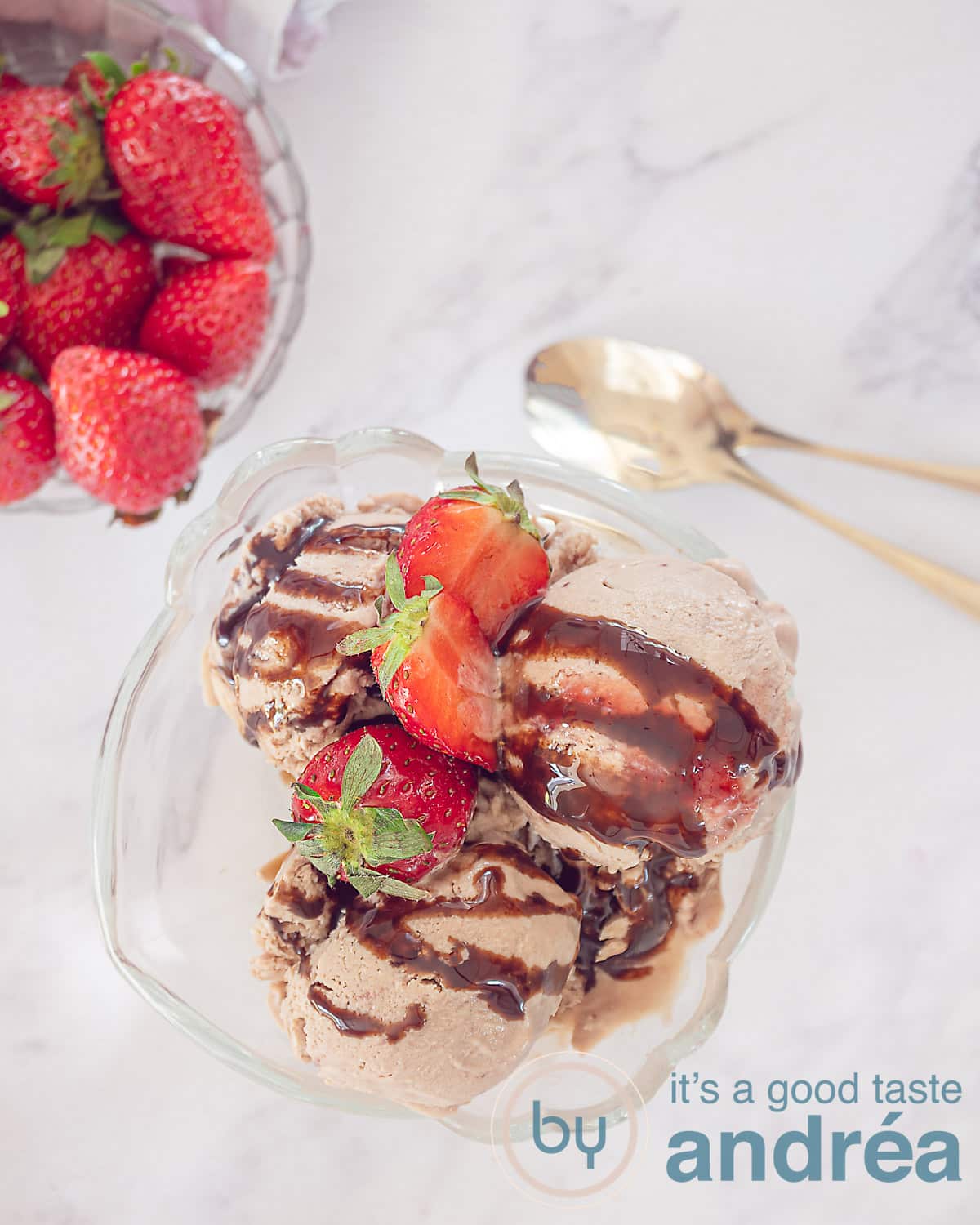 Een foto van bovenaf van een coupe gevuld met chocolade ijs van Griekse yoghurt, en aardbeien, verse aardbeien. Linksbovenin een glazen schaaltje met extra aardfbeien. Recht twee gouden  lepels. Een witte ondergrond.