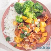 Een vierkante foto met een roze bord met drumsticks, bacon, mango, saus, geschaafde amandelen, rijst en broccoli