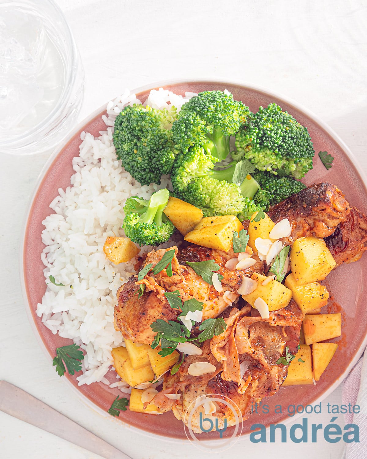 Een foto van bovenaf met een roze bord en een witte ondergrond. Op het bord liggen twee drumsticks met mango en saus, broccoli, geschaafde amandelen en rijst.