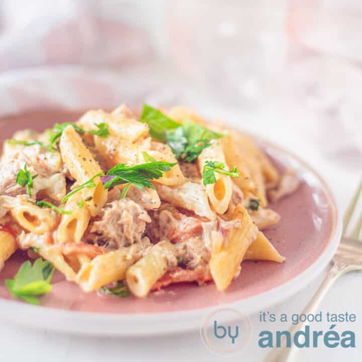 Een vierkante foto met een roze bord met pasta met cherrytomaten en tonijn en peterselie. Een roze achtergrond.