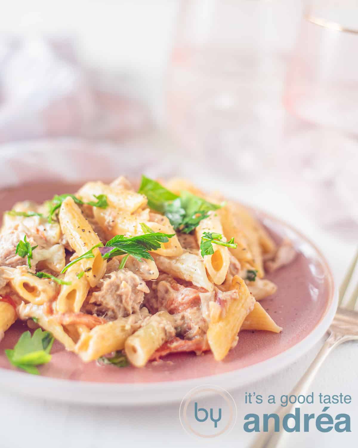 Een hoogtefoto met een deel van een roze bord met penne, tonijn en een romige saus. In de achtergrond twee roze glazen