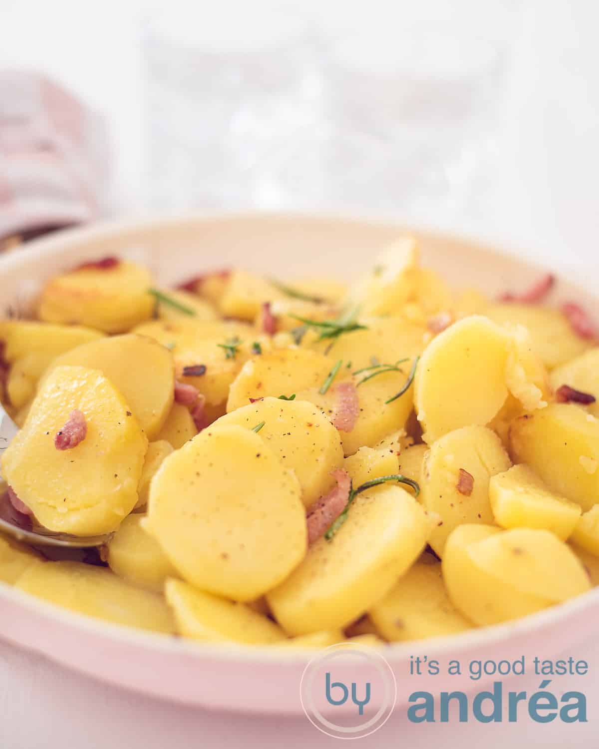 Een foto in de hoogte van een roze koekenpan met gebakken aardappels met spek, rozemarijn en ui. Een witte achtergrond.