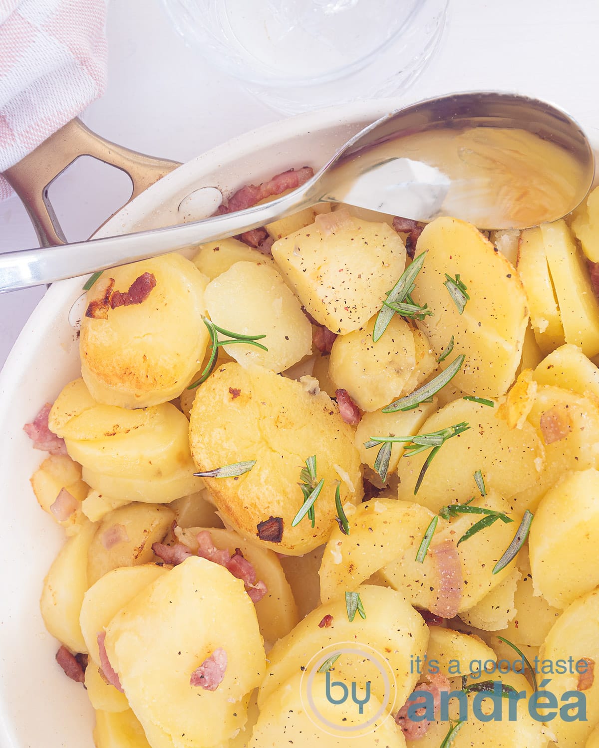 Een foto van bovenaf van een witte koekenpan met gebakken aardappels, ui, roemarijn en spekjes. Een lepel schept erin.