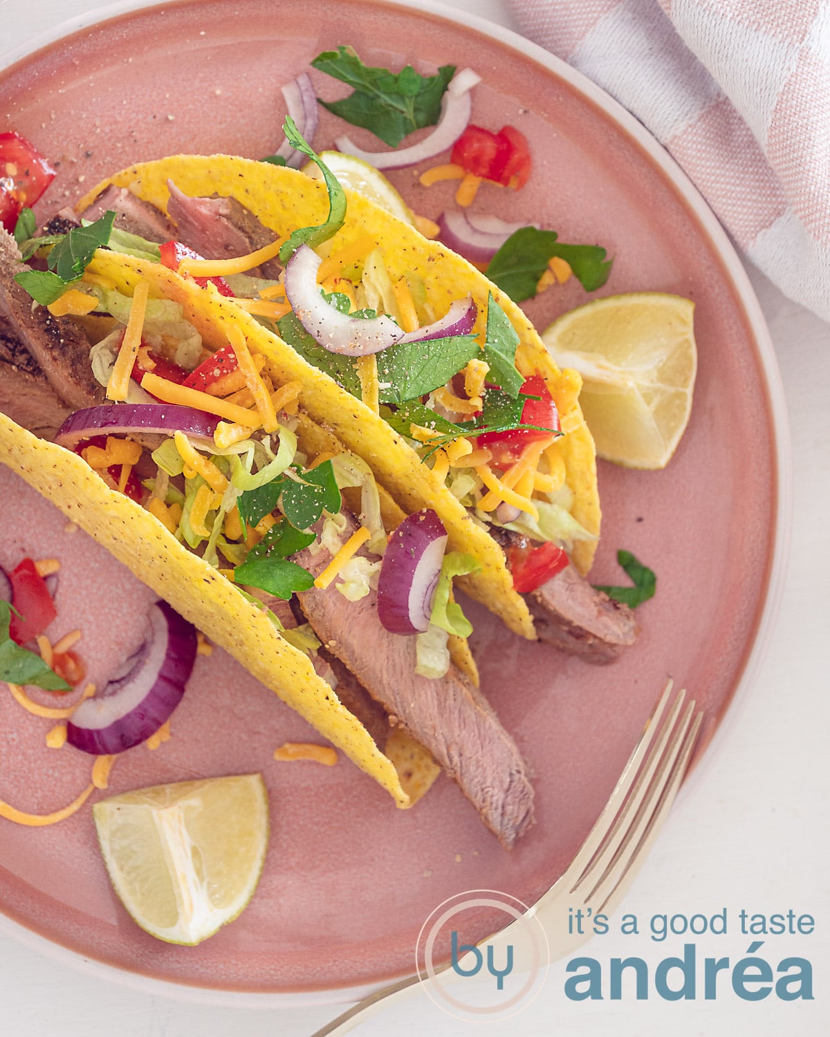 An overhead photo of a pink plate with two taco shells, filled with salad, onion, sirloin strips and cheese. Some garnish and lime wedges around it. A white background.