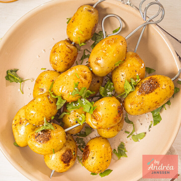 Bovenaanzicht van gekruide krieltjes op BBQ-spiesjes, geserveerd met grof zeezout en verse kruiden.