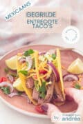Een roze bord met daarop tacoschelpen gevuld met gegrilde entrecote en salade. Limoenpartjes en cheddar kaas als garnering. Een textoverlay 