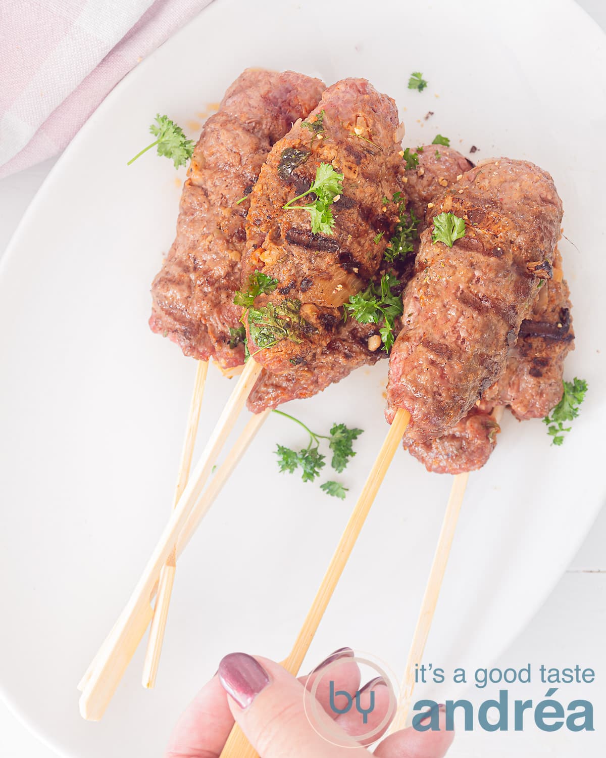 Een foto van bovenaf met spiesjes köfte kebab. Een hand pakt een spiesje. Op een witte schaal. 