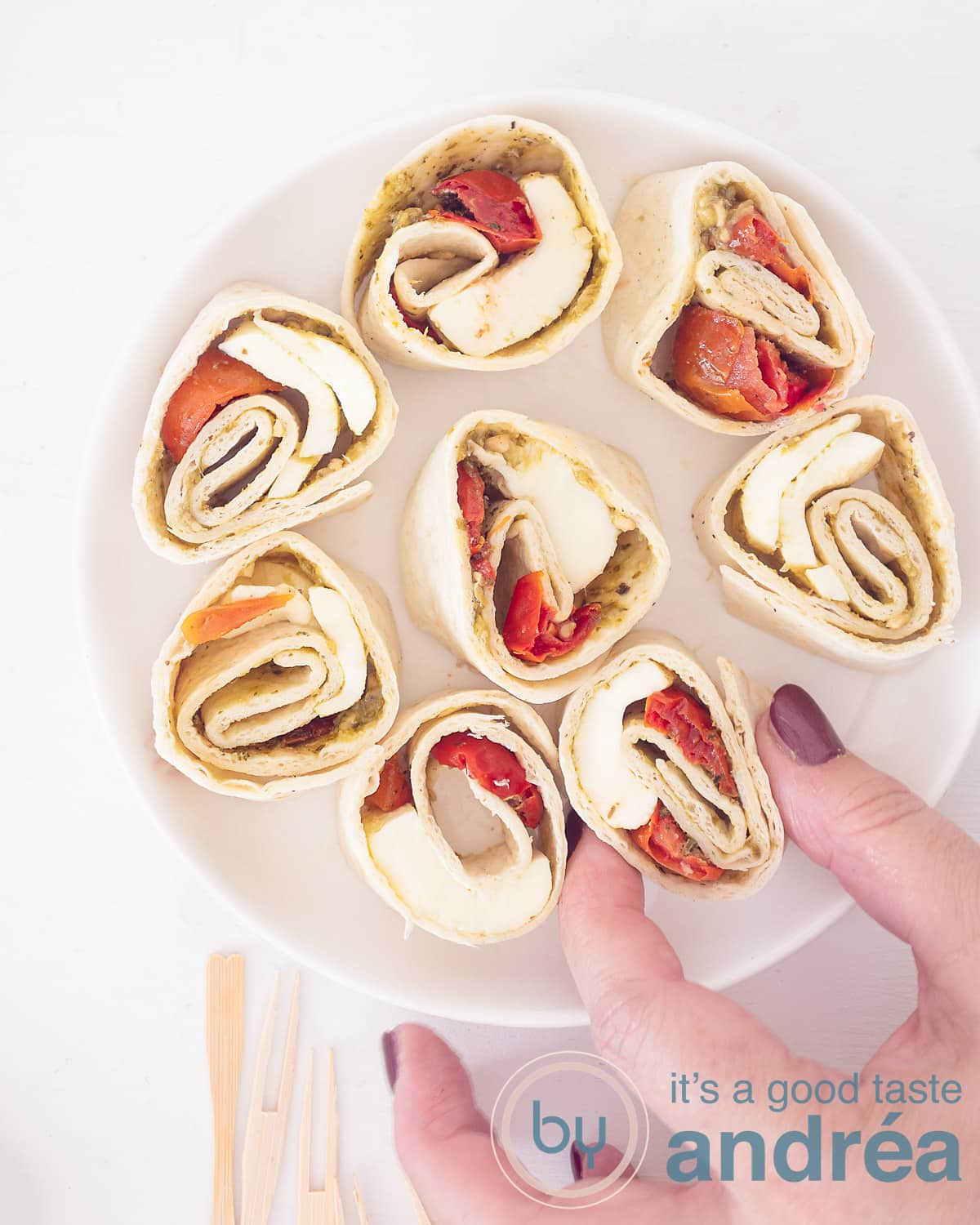 Een foto van bovenaf met acht Caprese wraps gevuld met tomaat, mozzarella en basilicum. Een hand pakt een hapje. Een witte ondergrond.