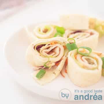 Een vierkante foto met een wit bord gevuld met ronde tortilla wrap snacks gevuld met kipfilet, kruiden roomkaas, komkommer en sla. Een witte achtergrond.