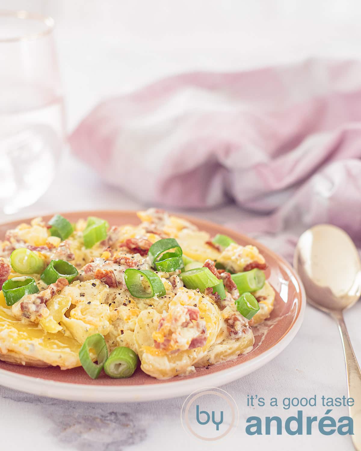 Een hoogtefoto met een roze bord gevuld met aardappelsalade met bacon en cheddar op een witte ondergrond. Een gouden lepel er rechts langs In de achterzijde een roze witte theedoek